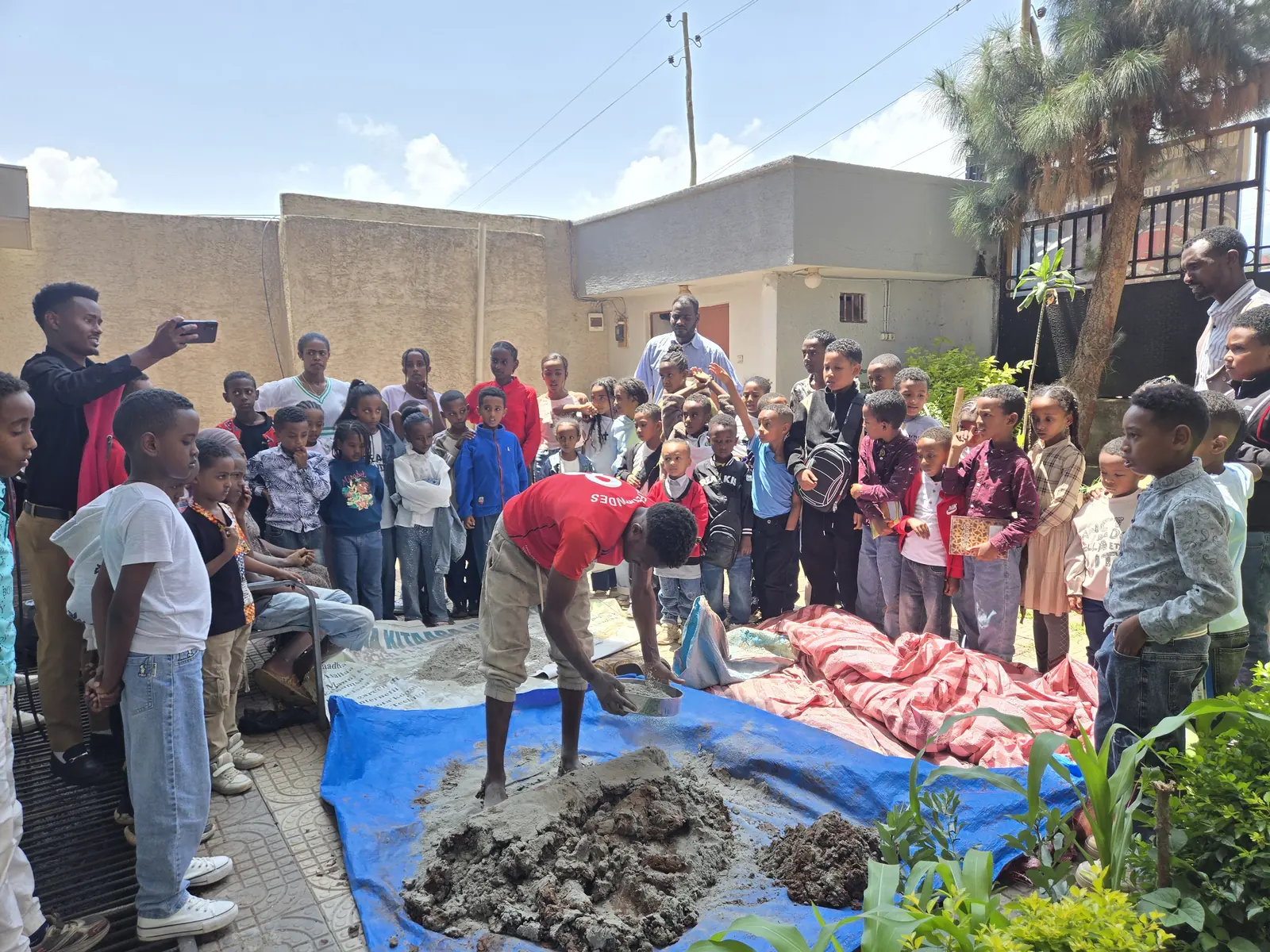 Children gather outdoors to watch a demonstration of preparing clay.