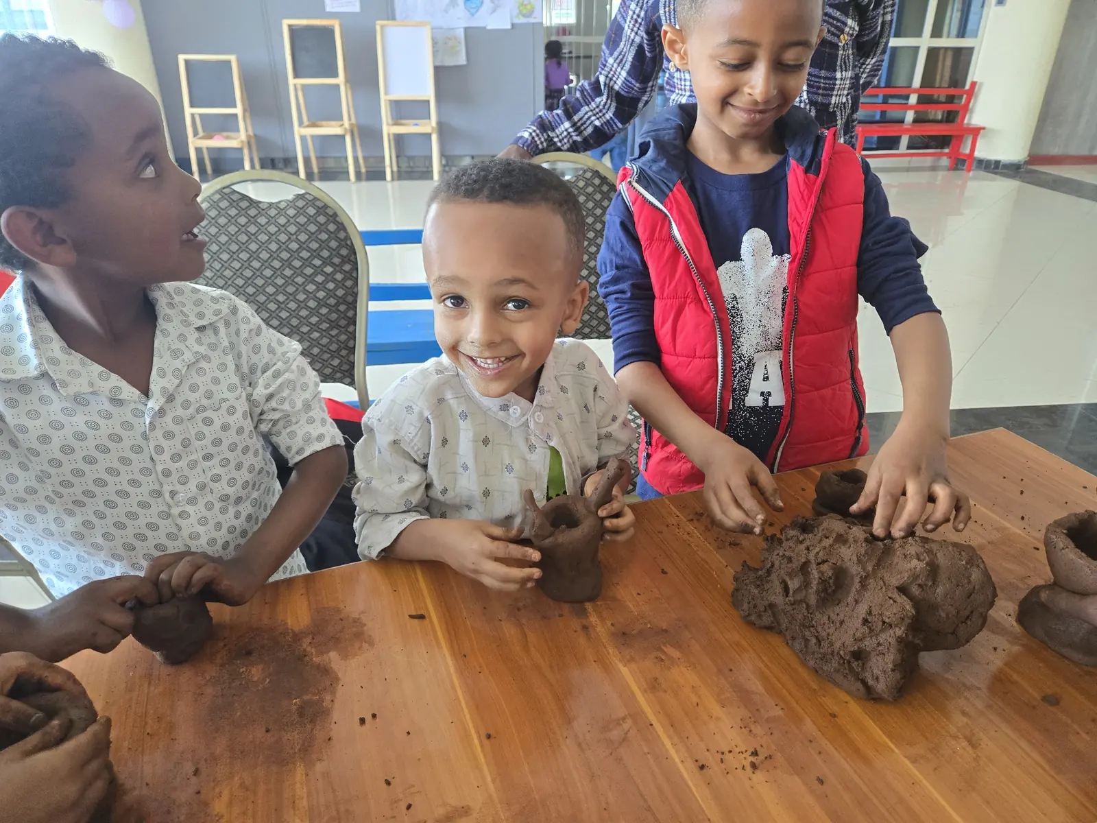 A smiling boy shapes a clay pot with friends at a wooden table.