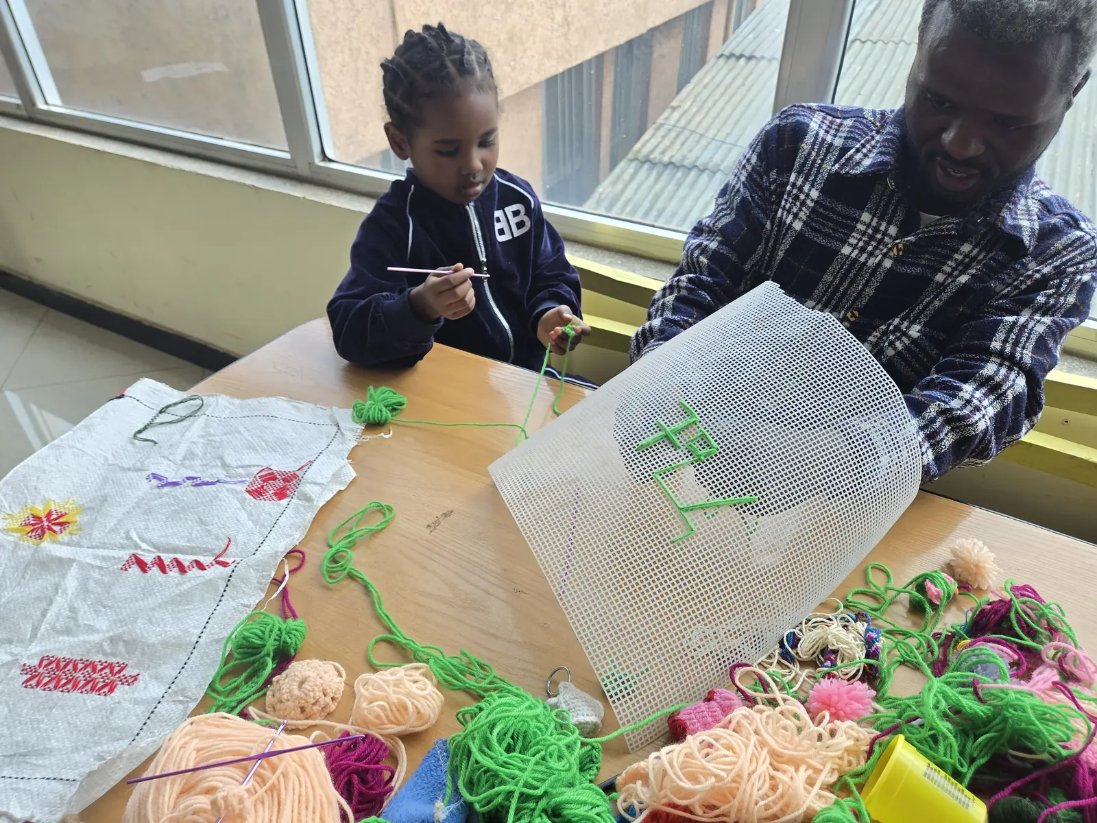 An adult helps a young girl stitch yarn into plastic canvas.