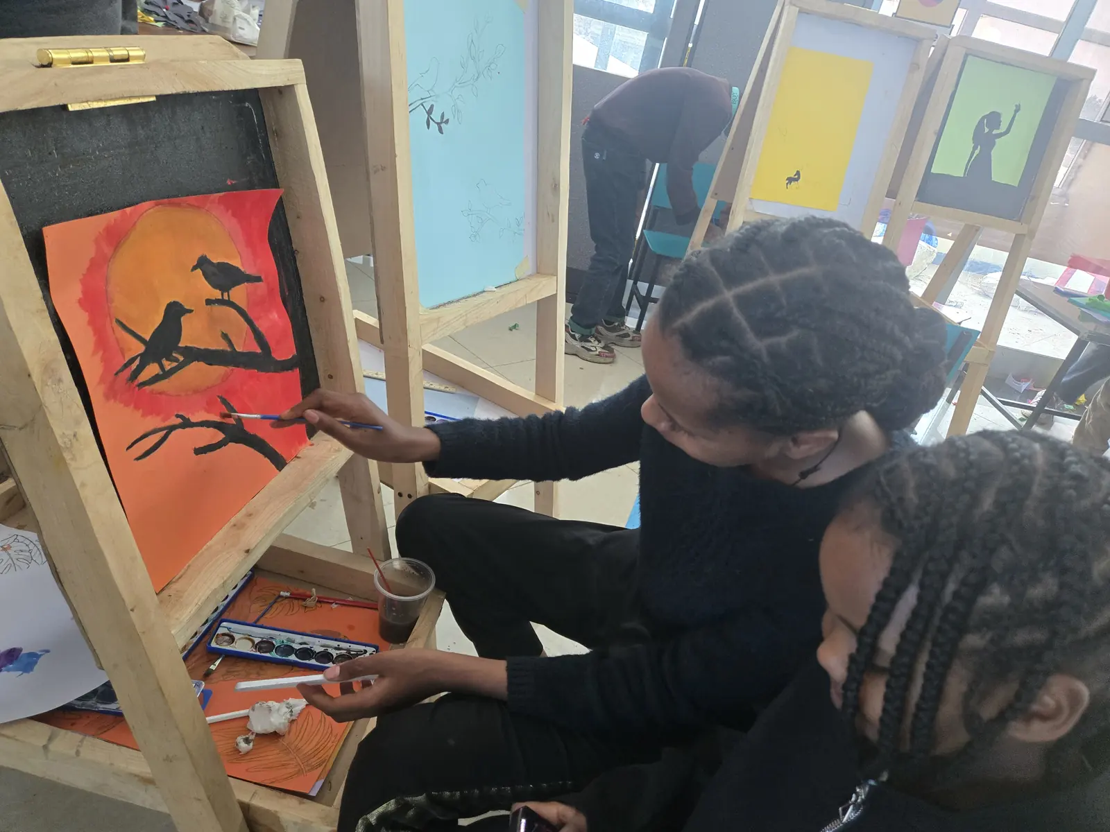 A girl paints birds on a branch against an orange sunset.