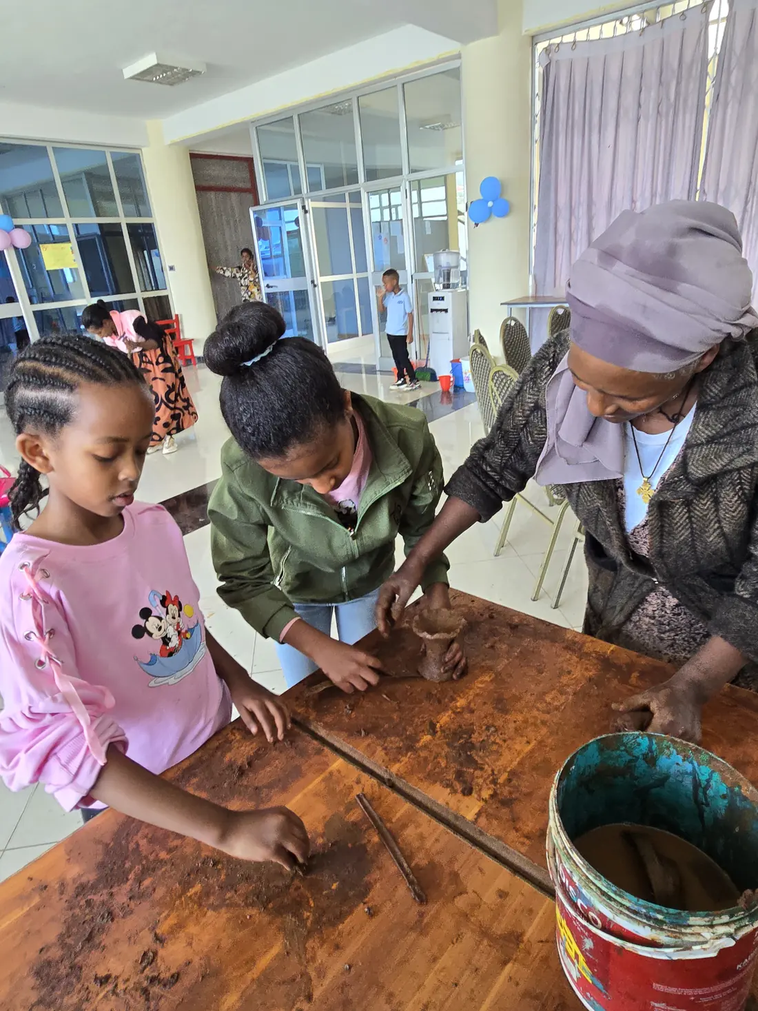 An elder shows two girls how to work clay by hand.