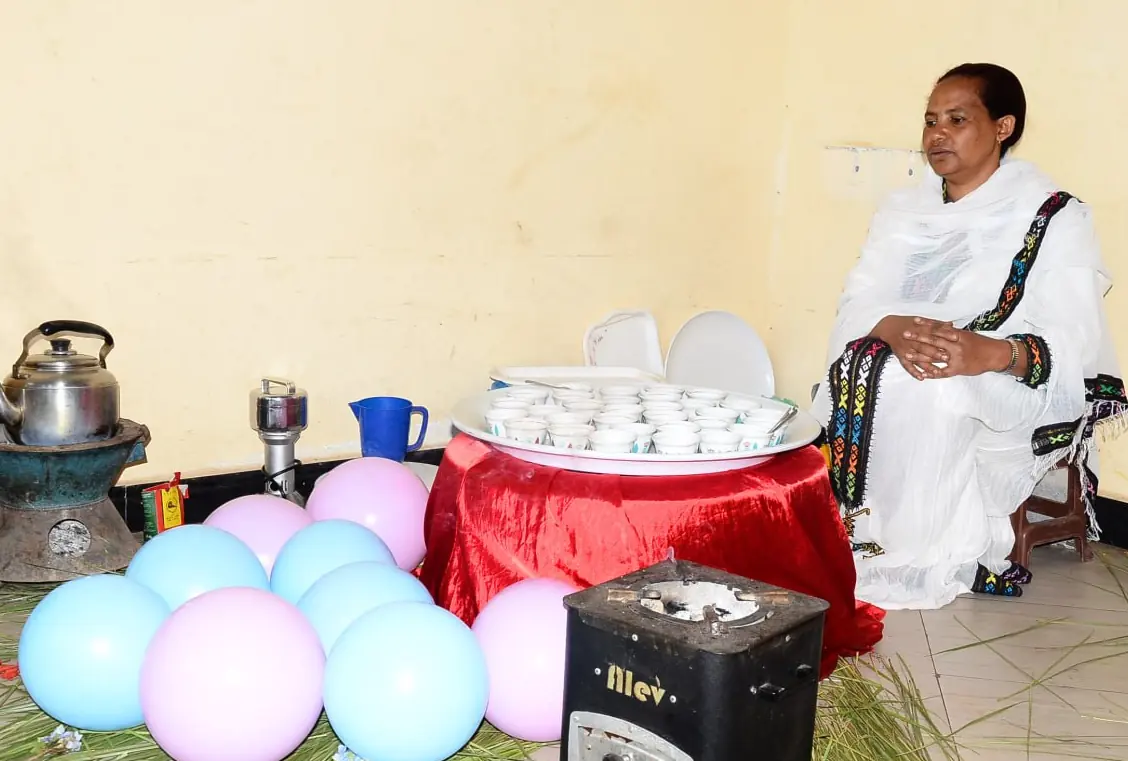 A woman in traditional dress prepares a coffee ceremony with a jebena, cups and balloons.