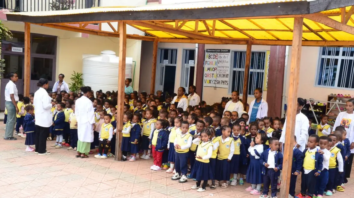 Kindergarten children stand in neat rows under the yellow canopy for assembly.