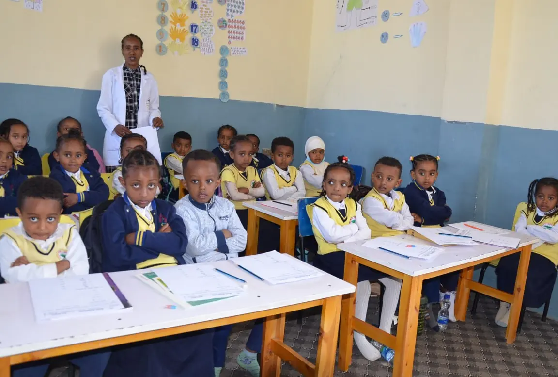A kindergarten class sits at small white tables, looking up at the camera.