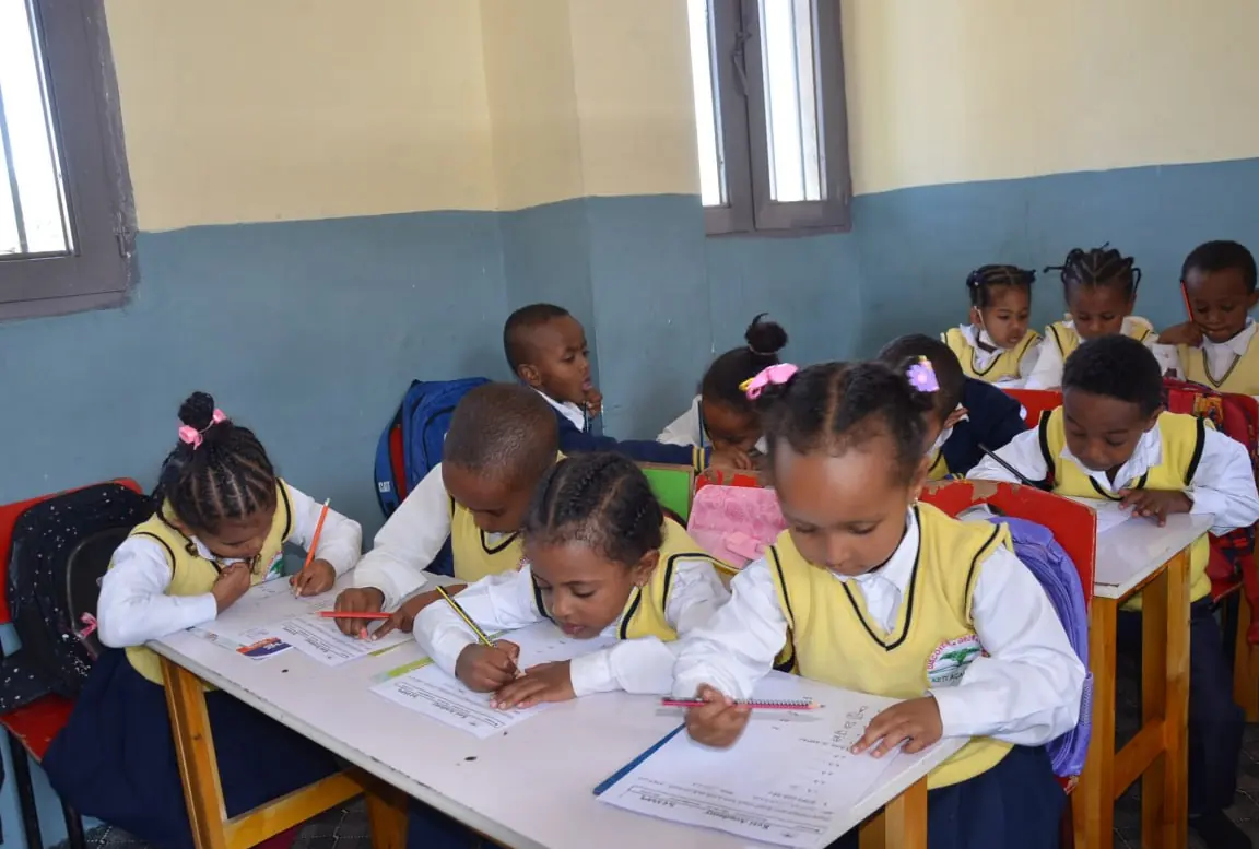 Kindergarten children colour worksheets at a shared table.