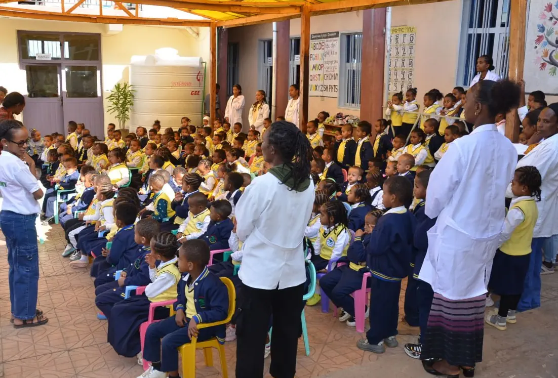 Teachers in white coats look after kindergarten children seated in the covered courtyard.