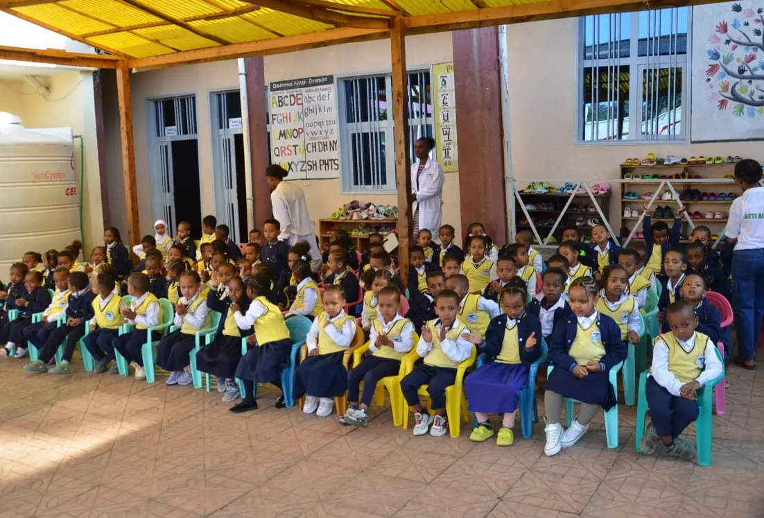 Kindergarten children in uniform sit in rows under the covered KG courtyard.