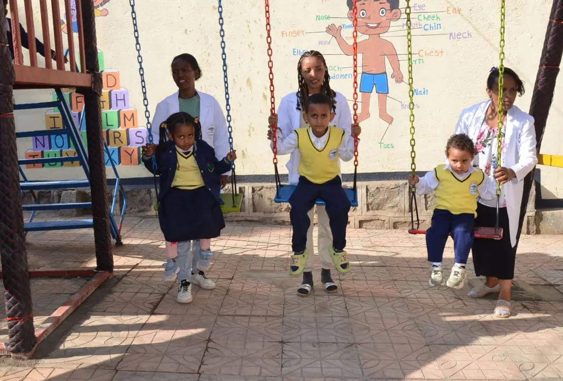 Three young children take turns on the swings, each with a teacher beside them.