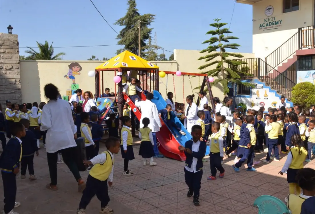 The KG yard full of children at play, with the school building and pine trees behind.