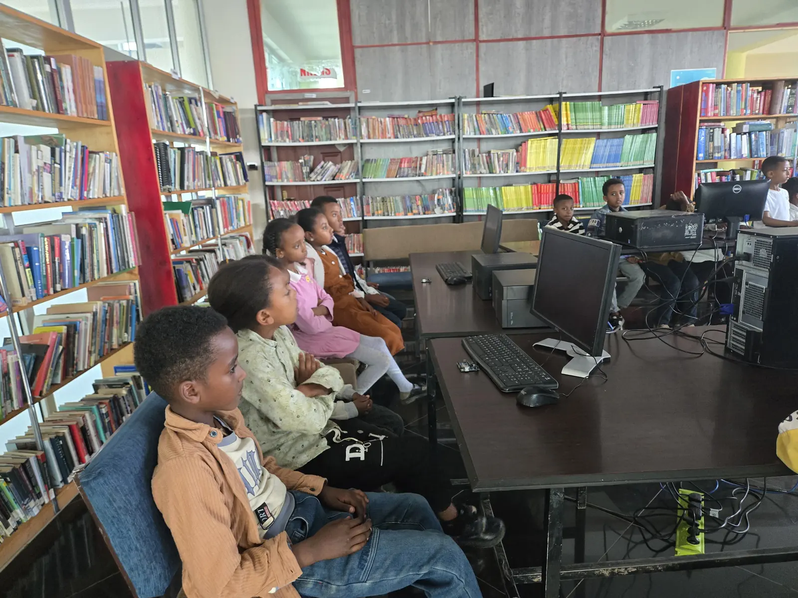 Children sit at computers among tall library bookshelves.