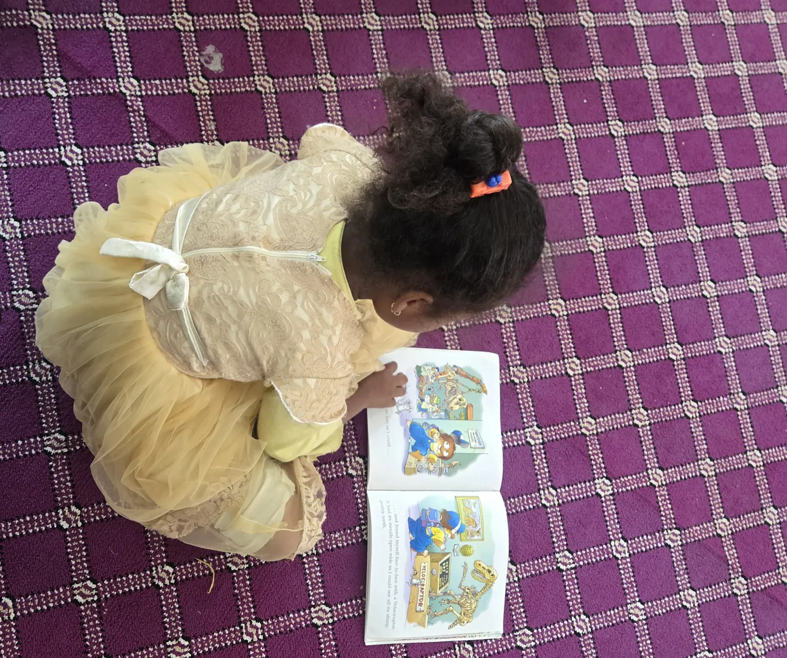 A girl in a yellow dress reads a picture book on the library carpet.