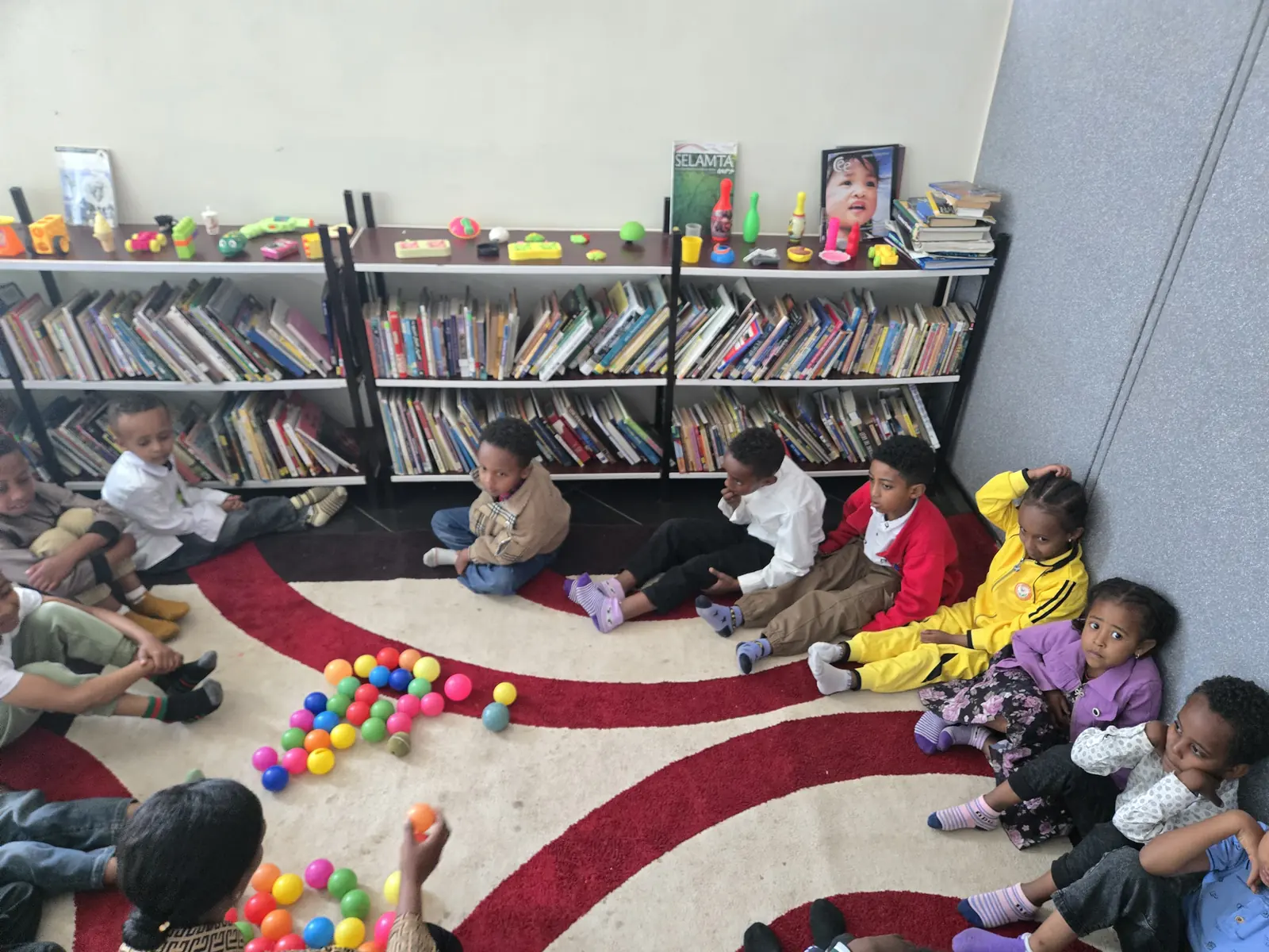 Young children sit on a rug in the children’s corner of the library, surrounded by books.