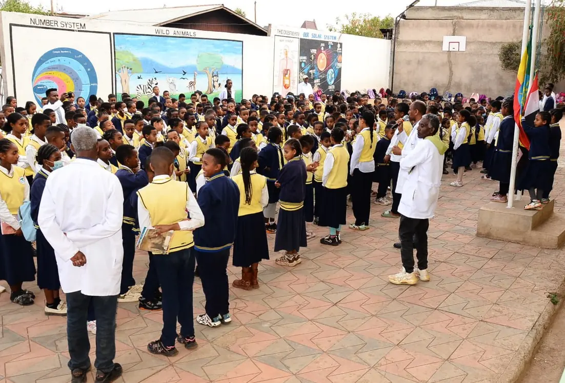 Students in navy and yellow uniforms gather for morning assembly beside the flagpole, with teachers in white coats.