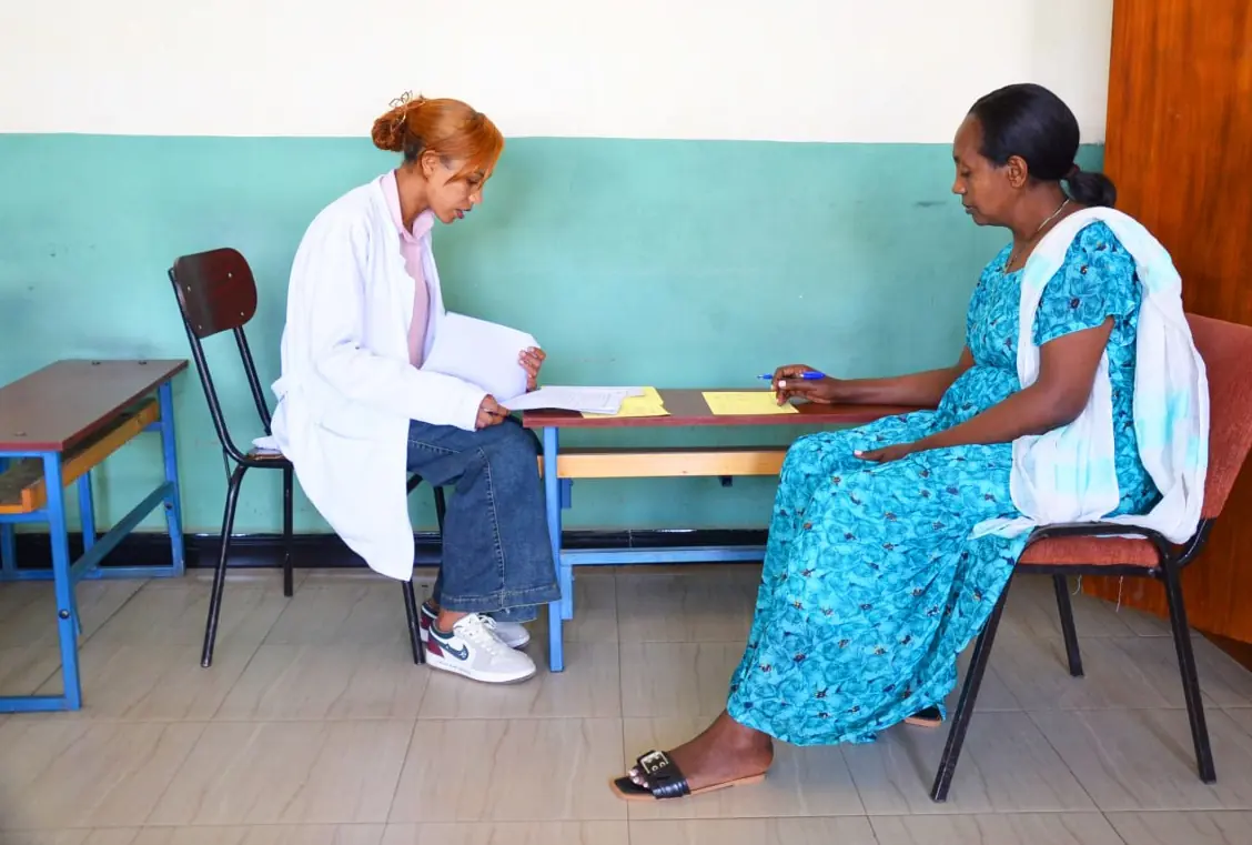 A teacher in a white coat meets a mother at a classroom desk during parent-teacher day.