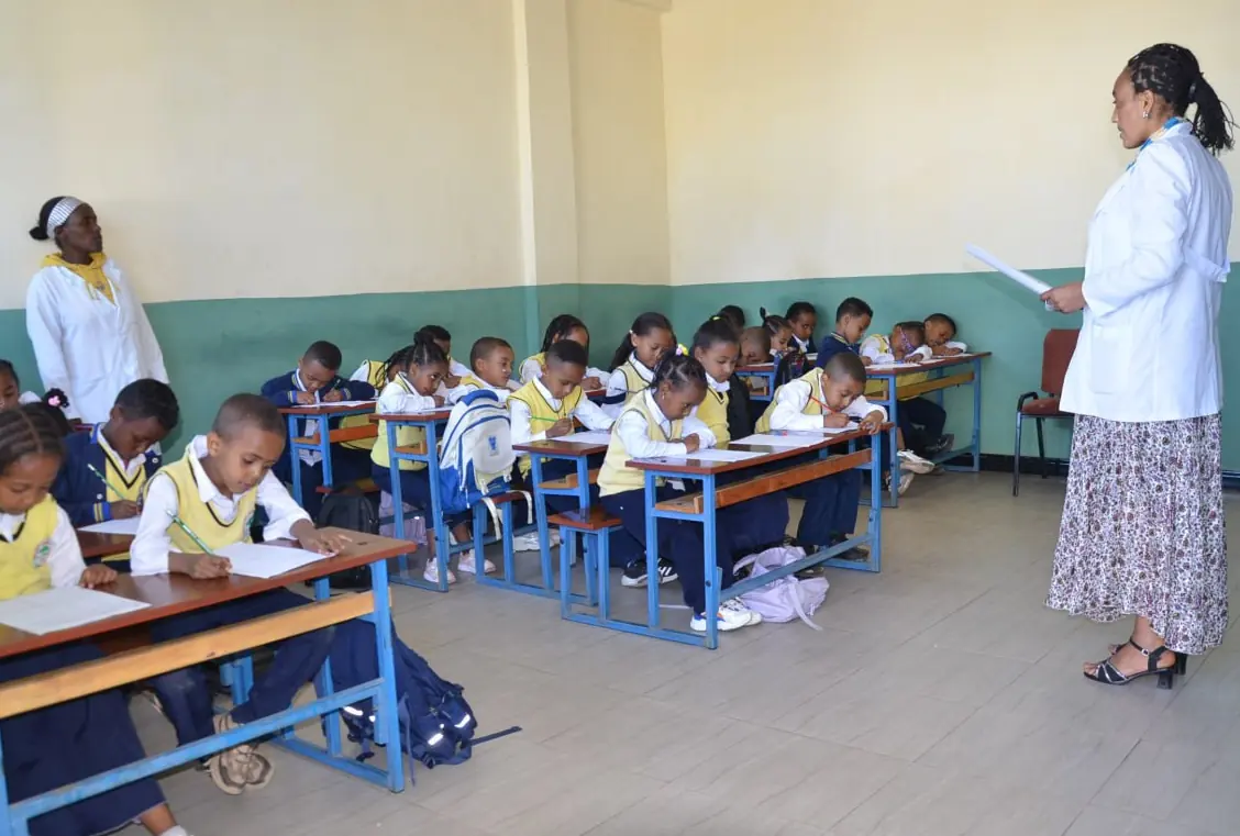 A teacher reads from a sheet while students write at rows of desks.