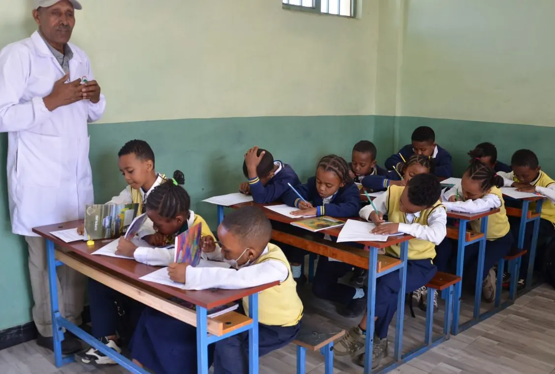A teacher in a white coat speaks to a class of primary students working at their desks.