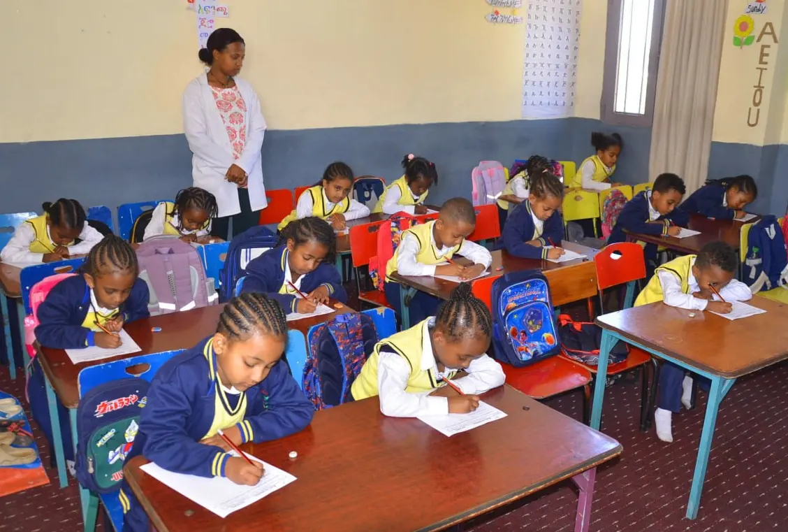First-grade students write at their desks, backpacks hung on their chairs.