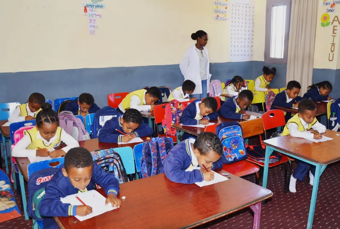 Young primary students at colourful desks with a teacher watching from the back.