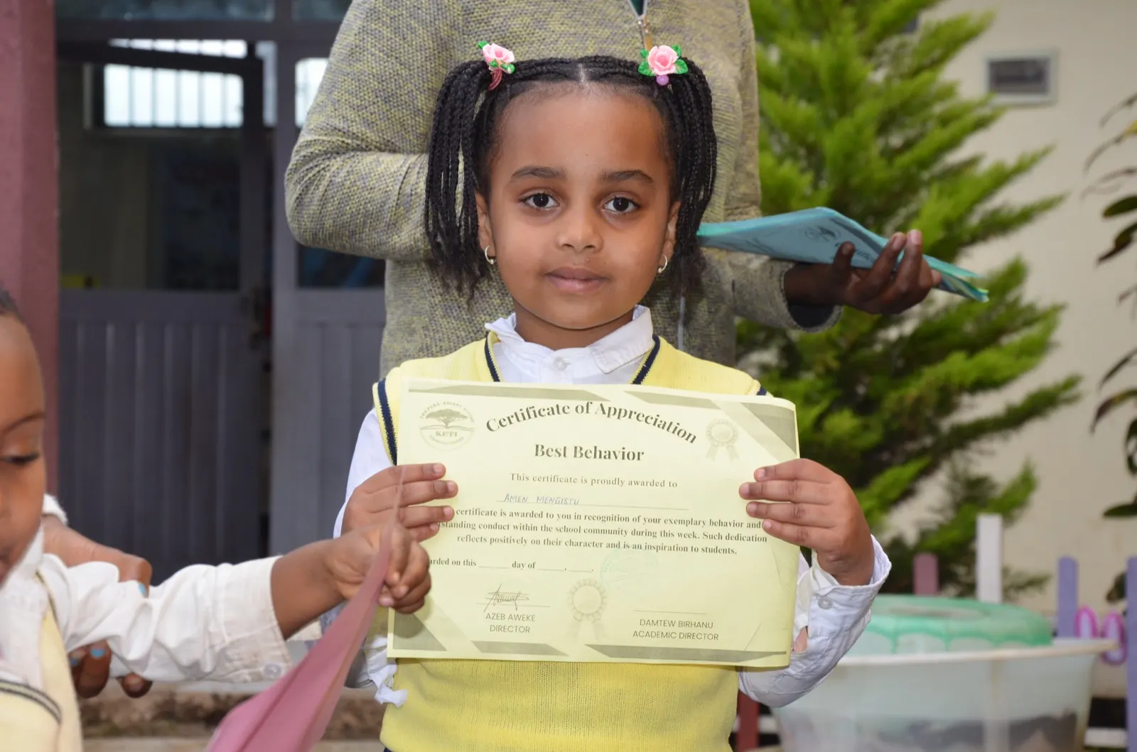 A Keti student holds a certificate of appreciation while a teacher stands behind her.